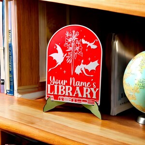 May include: Red wooden sign with a white dragon design and the text "Your Name's Library". The sign is on a wooden stand.