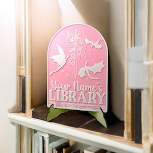 May include: Pink wooden sign with a white silhouette of a sword, flowers, and three dragons. The sign reads "Your Name's Library".