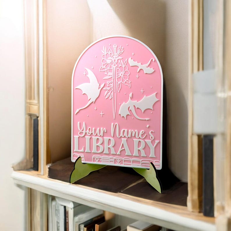 May include: Pink wooden sign with a white silhouette of a sword, flowers, and three dragons. The sign reads "Your Name's Library".