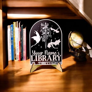 May include: Black wooden sign with a white design of a sword, flowers, and three dragons. The sign reads "Your Name's LIBRARY" and is on a wooden stand.