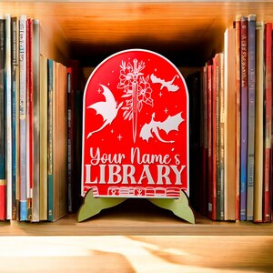 May include: Red wooden bookend with a white design of dragons and a sword. The bookend has a stand and says "Your Name's LIBRARY" in white letters.
