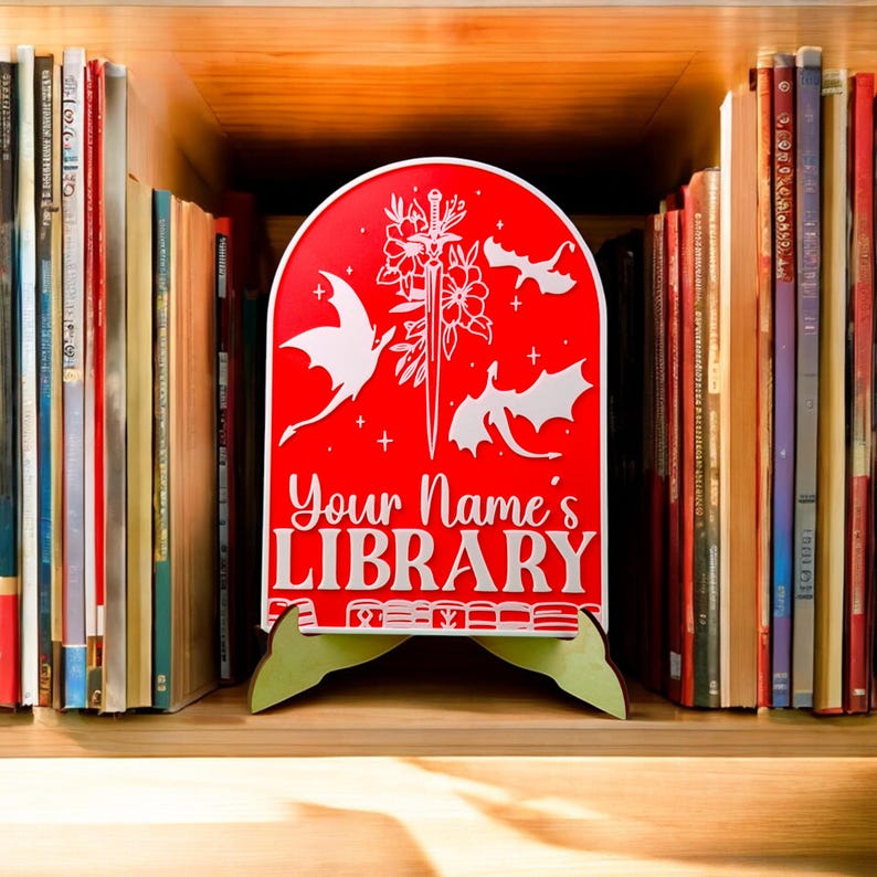 May include: Red wooden bookend with a white design of dragons and a sword. The bookend has a stand and says "Your Name's LIBRARY" in white letters.