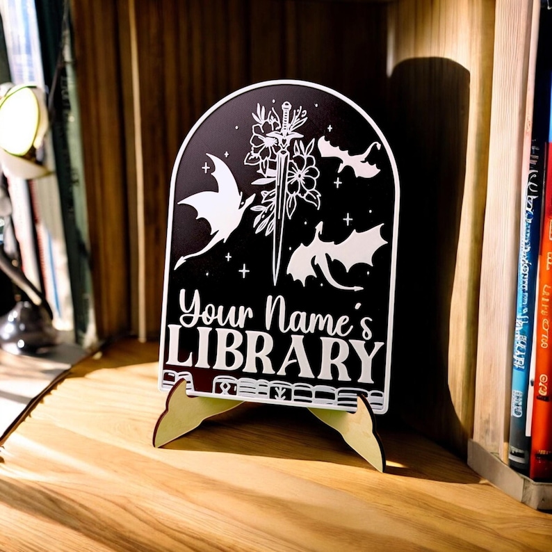 May include: Black and white wooden sign with a whimsical design featuring dragons, a sword, and flowers. The sign reads "Your Name's LIBRARY" and is on a small wooden stand.