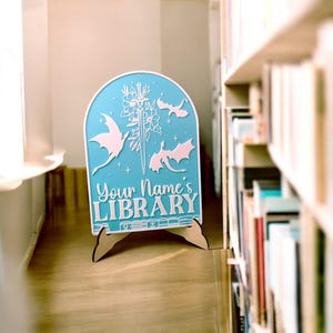 May include: A light blue wooden sign with a white silhouette of a sword, flowers, and dragons. The sign reads "Your Name's Library" and is on a wooden stand.