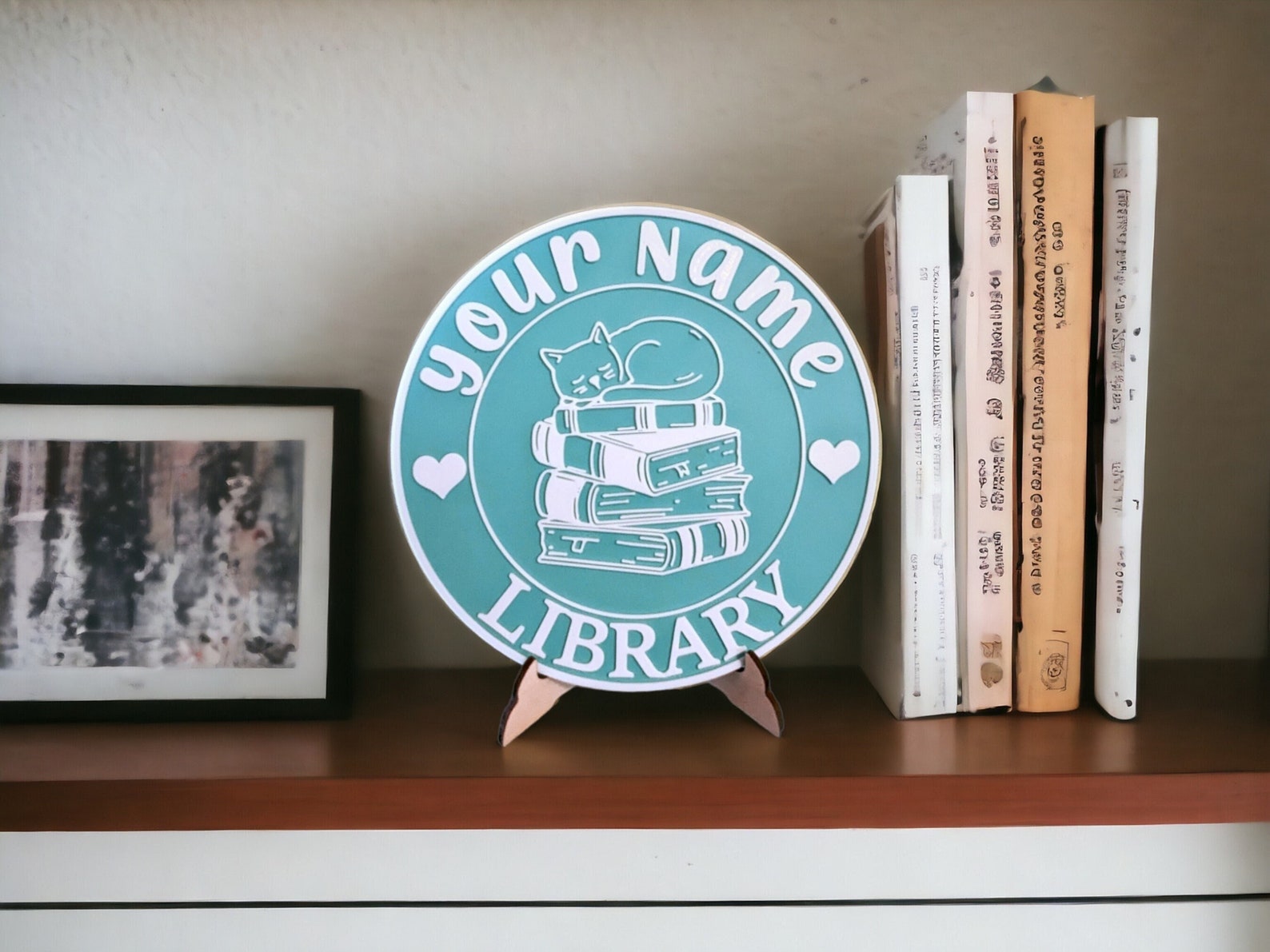 A round teal name plate rests on a wooden stand. The sign has a cat sleeping on a stack of books and reads "Your Name" on top and "Library" on the bottom.