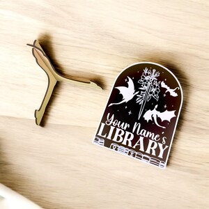May include: A black wooden sign with a white arch design featuring three dragons and a sword. The sign reads "Your Name's LIBRARY" with a stack of books at the bottom.
