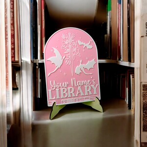 May include: Pink wooden sign with a white design of dragons and a sword. The sign reads "Your Name's Library".