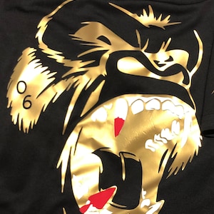 Puede incluir: Camiseta negra con un gráfico dorado de la cabeza de un simio rugiendo. El diseño incluye el número "06" y detalles rojos en los dientes. La camiseta está hecha de una tela suave y oscura.