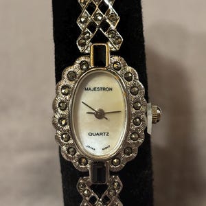 May include: A silver-toned Majestron quartz watch with a white oval face. The watch features a decorative bezel and band with small, dark-colored accents. The watch face displays the brand name, "Majestron," and the word "Quartz." The watch is on a black surface.