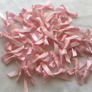 May include: A collection of small, light pink satin bows. The bows are tied and arranged in a pile, with the satin material reflecting light. The bows are likely for crafting, gift wrapping, or embellishments.