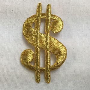 May include: A gold-colored dollar sign symbol patch featuring dense, metallic-looking embroidery thread. The design consists of the classic currency character with two vertical lines crossing through the center of the curved S-shaped form.