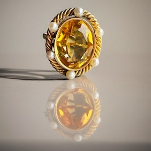 Puede incluir: Un anillo de oro con una gran piedra preciosa amarilla ovalada. El anillo está rodeado por un diseño de cuerda dorada trenzada y pequeñas perlas redondas. La reflexión del anillo es visible debajo.