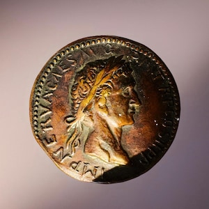 Puede incluir: Una moneda de bronce que presenta un perfil de una persona con una corona de laurel. La moneda tiene inscripciones detalladas alrededor del borde y está envejecida con una pátina oscura. El diseño de la moneda se destaca por la luz.