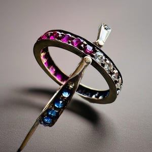 Könnte beinhalten: Ein silberner Ring mit einem Band aus abwechselnd rosa und klaren Edelsteinen. Der Ring hat ein einzigartiges Design mit einem vertikalen Balken, der mit blauen Edelsteinen besetzt ist, und einem kleinen, rechteckigen klaren Edelstein.