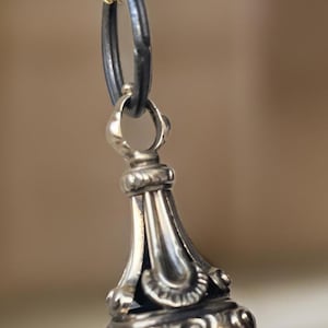 Puede incluir: Un colgante de sello de plata con detalles intrincados, colgado de una cadena negra con un cierre dorado.