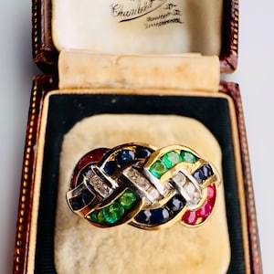 May include: A gold ring with a woven design, featuring alternating sections of red, blue, green, and white gemstones. The ring is set with diamonds and is displayed in a vintage jewelry box.