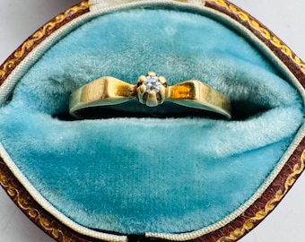 Könnte beinhalten: Ein goldener Ring mit einem kleinen, klaren Edelstein in einem blütenartigen Design. Der Ring wird in einer Vintage-Schmuckschatulle mit blauem Samtinnenraum und braunem Lederäußeren präsentiert. Die Schachtel ist mit "LONDON." gekennzeichnet.