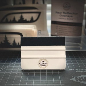 May include: White squeegee with a black felt edge and a logo that says "Accentrek Designs" with a mountain range inside a circle. The squeegee is on a gray cutting mat.