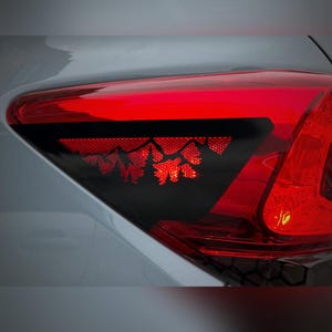 May include: A car's red tail light with a black section featuring a mountain and tree design. The light is bright red and triangular, with a white car body visible.