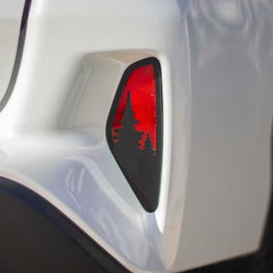 May include: A close-up of a car's white bumper with a black and red reflector. The reflector features a silhouette of pine trees against a red background. The design adds a decorative touch to the vehicle's exterior.