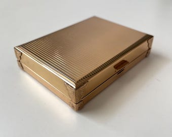 May include: A rectangular gold-colored compact case with a ribbed top and decorative corner accents. The compact has a small clasp on the side. The case is likely made of metal and is sitting on a white surface.