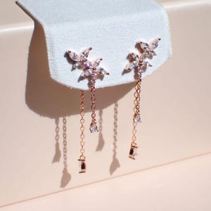 May include: A pair of rose gold earrings with a delicate floral design and dangling chains accented with clear cubic zirconia stones.