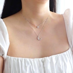 Puede incluir: Un collar de plata con un colgante de media luna y un pequeño colgante redondo. El collar está en capas y tiene una cadena delicada.