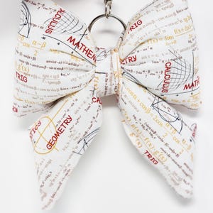 May include: A white fabric bow-tie keychain printed with mathematical equations and terms like "Calculus," "Geometry," and "Trig." The bow is attached to a silver-colored metal clasp.