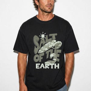 Könnte beinhalten: Ein schwarzes Kurzarm-T-Shirt mit einem Grafikdesign, das den Text SALT OF THE EARTH und MATTHEW 5:13 zeigt. Das Design enthält eine Surfbrett-Illustration, ein Sonnensymbol und stilisierte Meereswellen in gedeckten Grau- und Weißtönen.