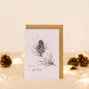 Op de afbeelding: Een witte wenskaart met een bruine envelop toont een gedetailleerde zwart-wit illustratie van een dennenappel en naalden. De woorden "Seasons Greetings" zijn in cursief geschreven. De kaart staat tegen een zachte, neutrale achtergrond.