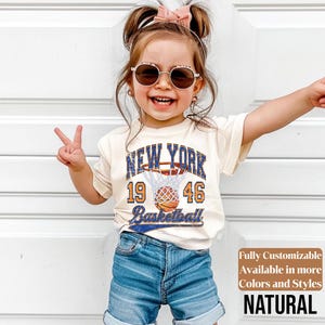 May include: A young child wearing a cream-colored t-shirt with a New York basketball graphic. The shirt features the text "NEW YORK 19 46 Basketball" in blue and orange. The child is also wearing denim shorts and sunglasses.