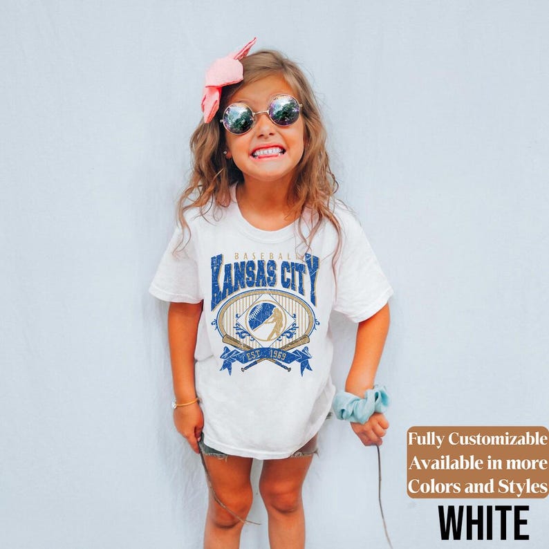 May include: White t-shirt with a blue and gold graphic that says "Kansas City Baseball" with a baseball and crossed bats. The shirt is worn by a young person wearing sunglasses and a pink bow.