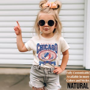 May include: A cream-colored t-shirt featuring a vintage-style baseball graphic with the text "Baseball Chicago" in blue and red. The design includes a baseball, crossed bats, and a banner. The shirt is paired with gray denim shorts.
