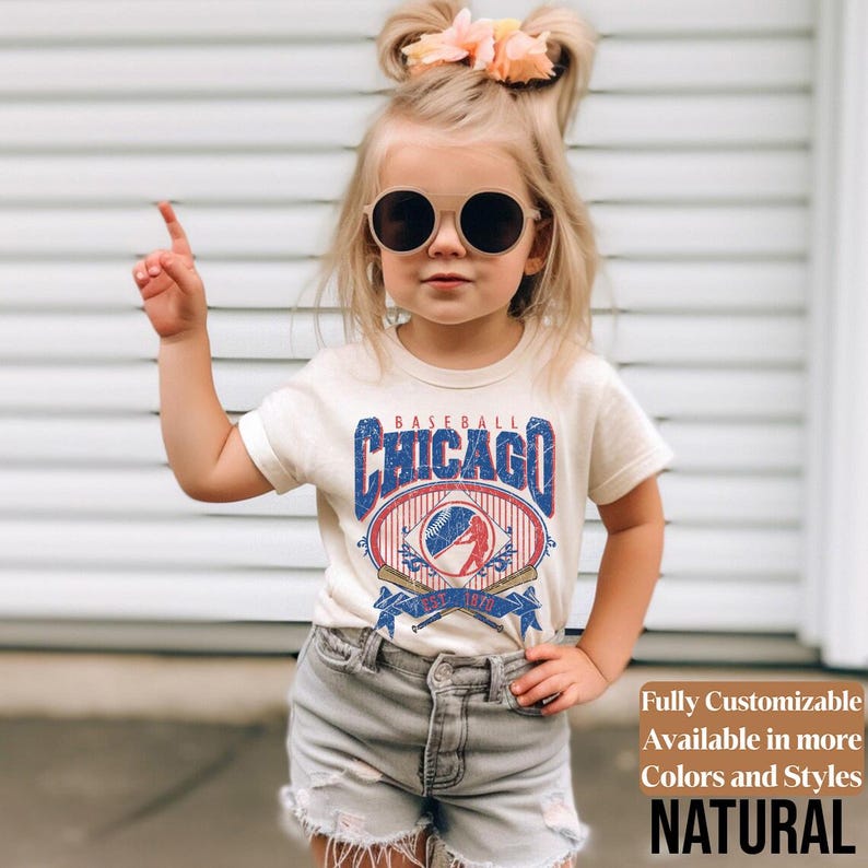 May include: A cream-colored t-shirt featuring a vintage-style baseball graphic with the text "Baseball Chicago" in blue and red. The design includes a baseball, crossed bats, and a banner. The shirt is paired with gray denim shorts.