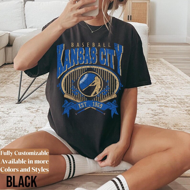 May include: Black t-shirt with a vintage-style baseball graphic. The design features the text "Kansas City" in blue, an oval with a baseball and batter, and "EST. 1969." The shirt is a dark color.