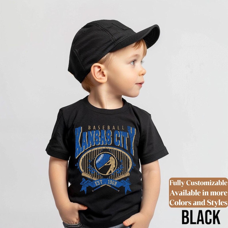 May include: A black t-shirt featuring a baseball-themed graphic with the text "Kansas City" in blue and gold. The shirt includes the text "EST. 1969" and a baseball bat design. The image also shows a black cap.