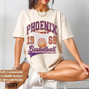 Peut inclure: T-shirt ivoire avec un motif de basket-ball Phoenix. Le motif comprend le mot "PHOENIX", les chiffres "19" et "68" et le mot "Basketball". Le graphisme est violet et orange.
