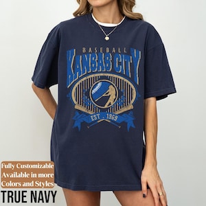 May include: Navy blue baseball t-shirt with a distressed graphic design. The design features the text "BASEBALL KANSAS CITY" above an oval emblem with a baseball player silhouette, baseball, and crossed bats. The text "EST. 1969" is also included.