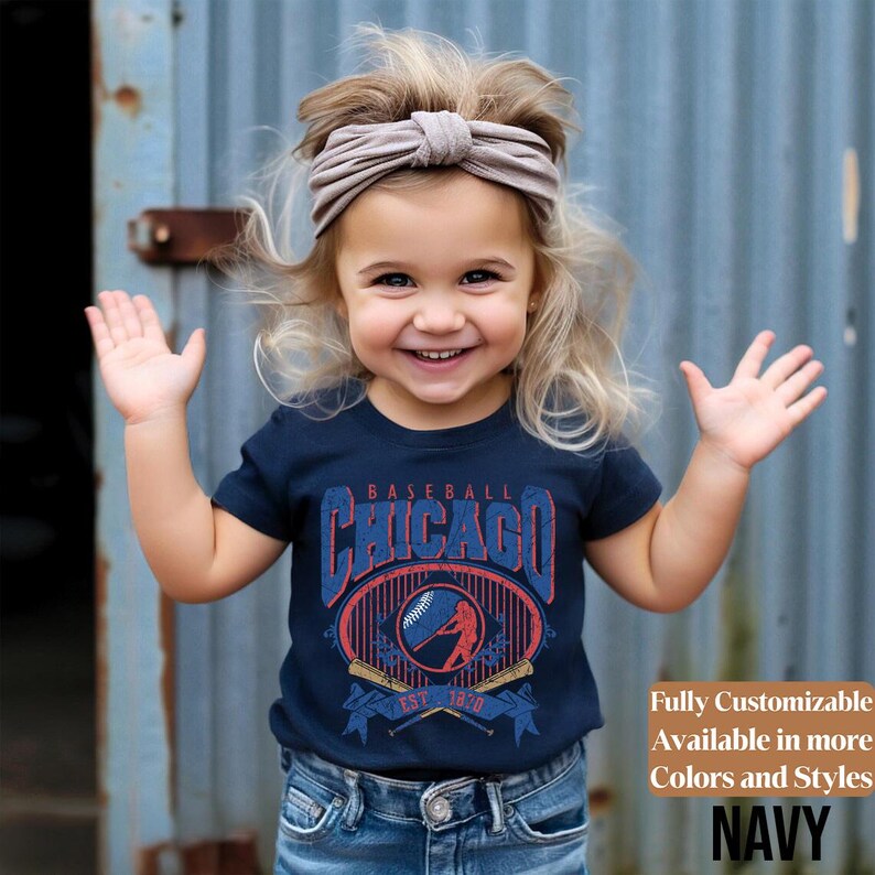 May include: A young child wearing a navy blue t-shirt with a Chicago baseball design. The shirt features the word "Chicago" in a vintage font, a baseball, and crossed bats. The text "EST. 1920" is also visible. The child is also wearing blue jeans and a beige headband.