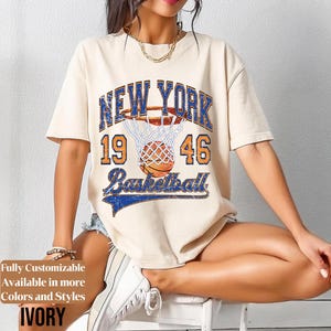 May include: Ivory-colored t-shirt with a distressed graphic featuring "NEW YORK 1946 Basketball" in blue and orange. The design includes a basketball going into a net. The shirt has short sleeves and a relaxed fit.