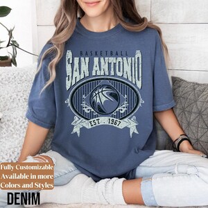 Może przedstawiać: Niebieska koszulka dżinsowa z postarzaną grafiką z napisem "BASKETBALL SAN ANTONIO EST. 1967". Grafika przedstawia koszyk&oacute;wkę w owalnej ramce. Koszulka jest z kr&oacute;tkim rękawem i okrągłym dekoltem.