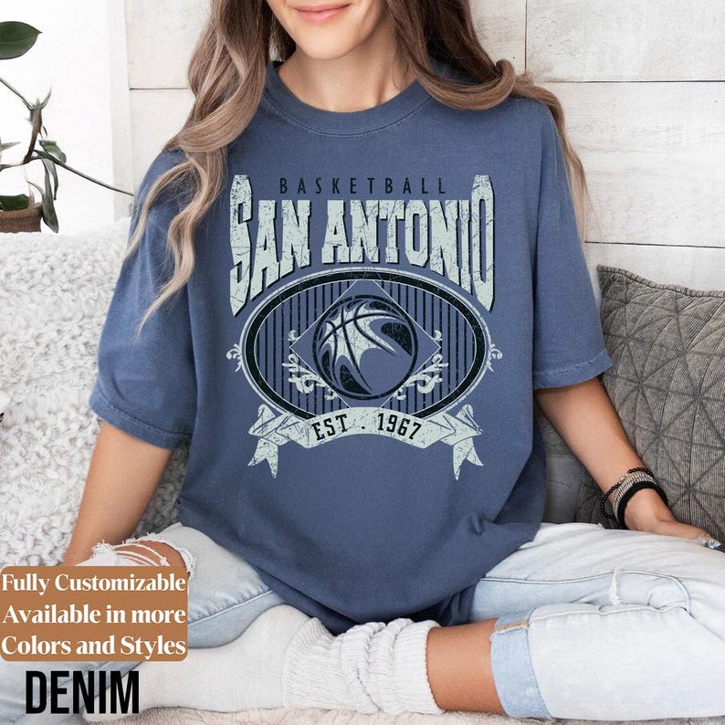 Może przedstawiać: Niebieska koszulka dżinsowa z postarzaną grafiką z napisem "BASKETBALL SAN ANTONIO EST. 1967". Grafika przedstawia koszyk&oacute;wkę w owalnej ramce. Koszulka jest z kr&oacute;tkim rękawem i okrągłym dekoltem.