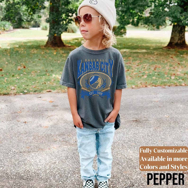 May include: A gray t-shirt with a blue and gold graphic that says "Baseball Kansas City Est. 1969". The shirt is paired with light blue jeans and black and white checkered shoes. The image also includes text that says "Fully Customizable Available in more Colors and Styles PEPPER".