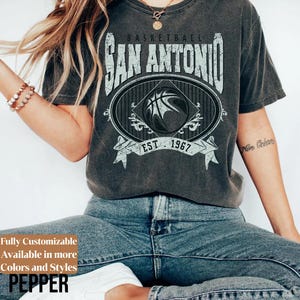 Może przedstawiać: Ciemnoszara koszulka z wyblakłą grafiką koszyk&oacute;wki San Antonio. Projekt zawiera napis "BASKETBALL SAN ANTONIO EST 1967" oraz grafikę koszykarską. Część linii odzieży z możliwością personalizacji.