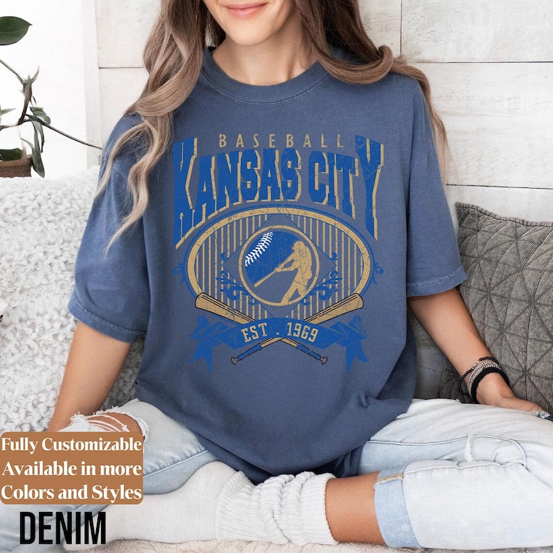 May include: Denim blue t-shirt with a distressed baseball graphic. The graphic features the text "Kansas City" in blue, a baseball, a batter silhouette, and crossed bats. The shirt also has the text "EST. 1969".