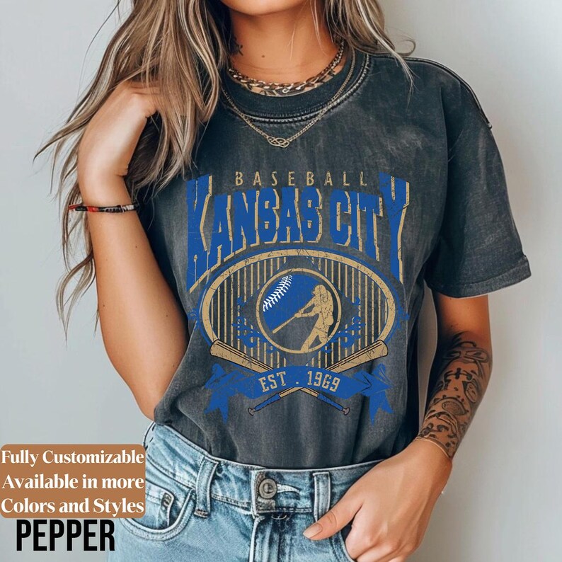 May include: Dark gray t-shirt featuring a blue and gold baseball design with the text "Kansas City" and "Est. 1969". The shirt has a vintage look and includes the word "Pepper" at the bottom.