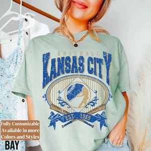 May include: A light green t-shirt with a distressed graphic featuring "Kansas City Baseball" in blue. The design includes a baseball, batter silhouette, and crossed bats, with "EST. 1969" on a blue banner. Text on the left says "Fully Customizable Available in more Colors and Styles BAY".