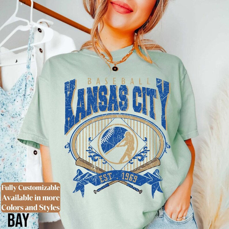 May include: A light green t-shirt with a distressed graphic featuring "Kansas City Baseball" in blue. The design includes a baseball, batter silhouette, and crossed bats, with "EST. 1969" on a blue banner. Text on the left says "Fully Customizable Available in more Colors and Styles BAY".