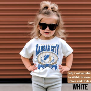 May include: A white t-shirt with a blue and gold baseball-themed graphic that says "Kansas City" and "Est. 1969". The shirt is worn by a young child wearing sunglasses. The text "Fully Customizable Available in more Colors and Styles WHITE" is also visible.