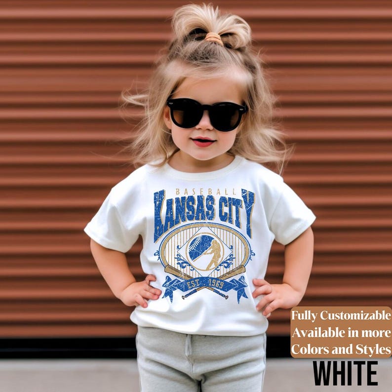 May include: A white t-shirt with a blue and gold baseball-themed graphic that says "Kansas City" and "Est. 1969". The shirt is worn by a young child wearing sunglasses. The text "Fully Customizable Available in more Colors and Styles WHITE" is also visible.